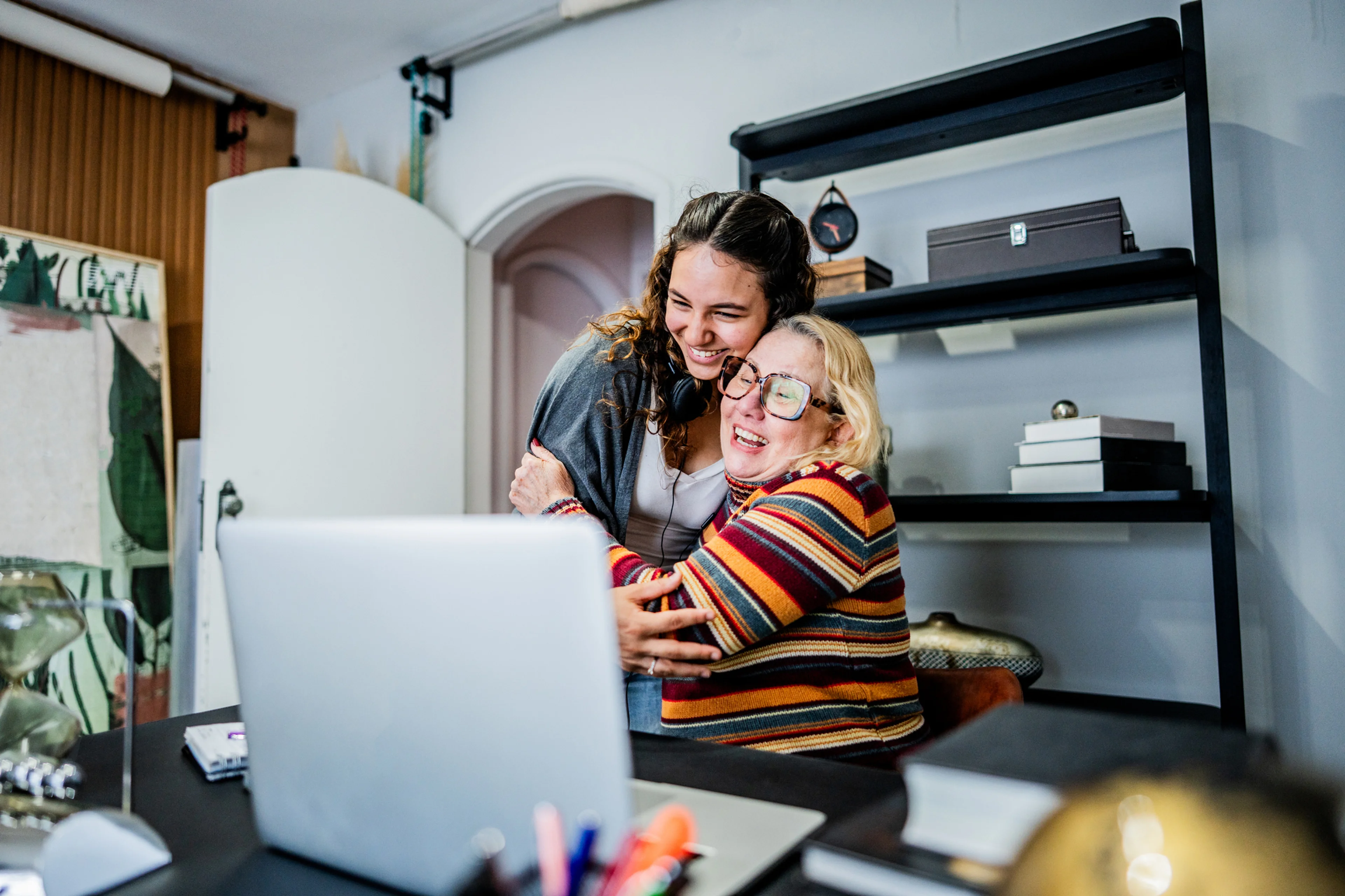 A heartwarming scene of two women sharing an intimate, joyful moment together while working on a laptop. Their close physical embrace and bright smiles capture the deep emotional connection and support between them, set in a cozy home office environment.